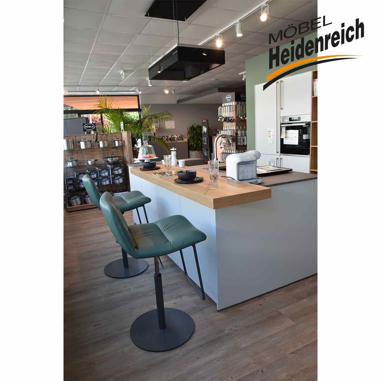 Blick auf die Kochinsel, Holzplatte darauf mit Kaffeetassen und Gläsern, davor zwei Barhocker.