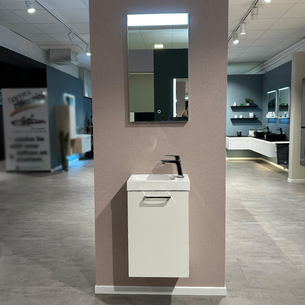 Badmöbel für das Gäste-WC. Oben ein beleuchteter Spiegel, unten in weiß Waschbecken mit Chrom Armatur und kleinem schmalem Unterschrank mit Tür.